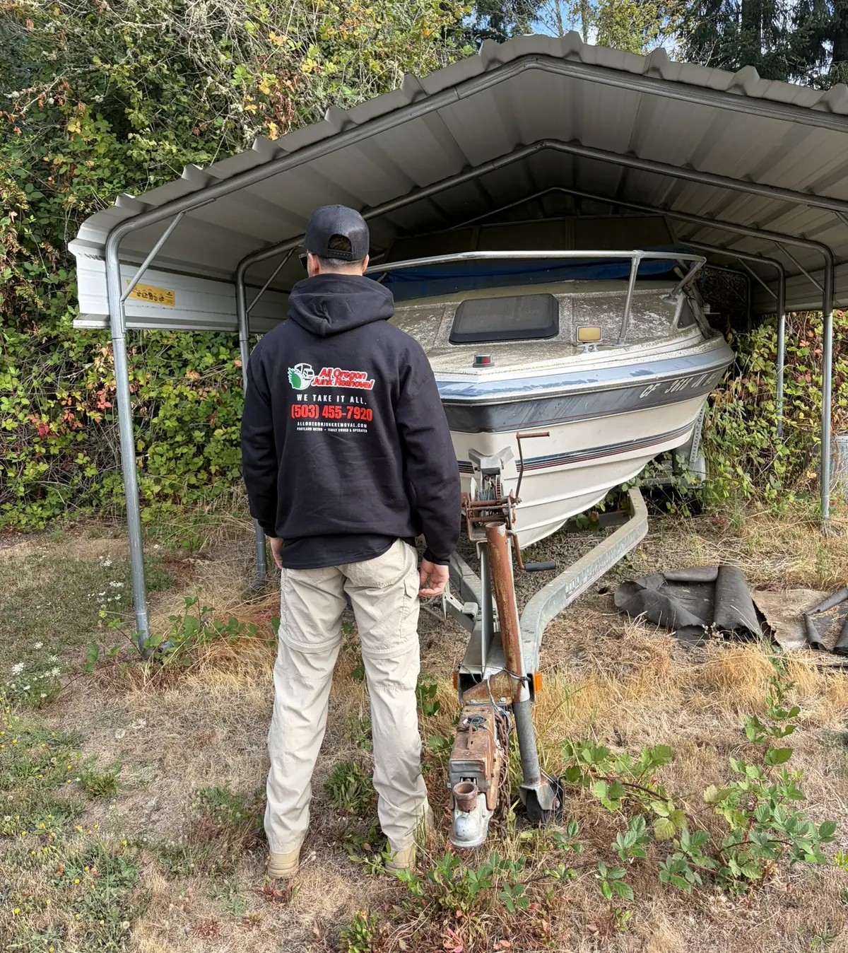 All Oregon Junk Removal crew member in a branded hoodie assessing a boat for removal from a carport