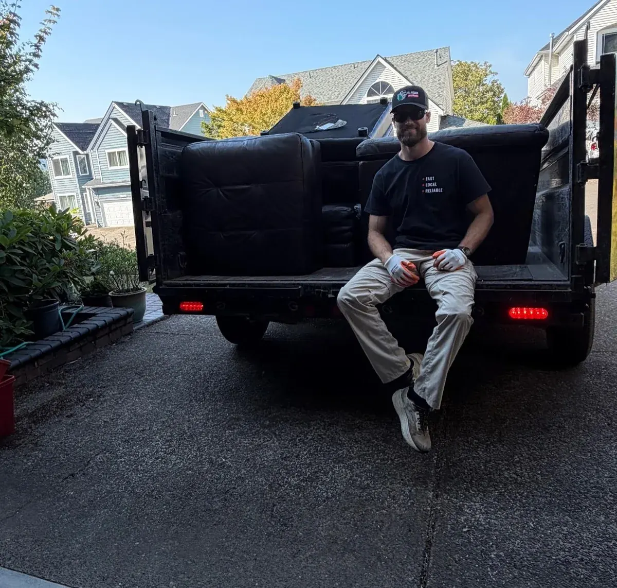 All Oregon Junk Removal crew member sitting on a loaded dump trailer holding an old couch, in a Portland-area driveway
